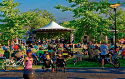Crowd of families sitting in lawn chairs and on the grass at an outdoor park pavilion, watching a live band perform at dusk, with bicycles parked in the foreground.