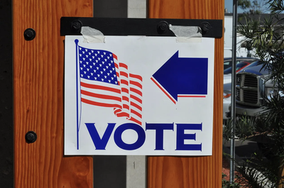 A "Vote" sign with an American flag and a blue arrow pointing left, taped to a wooden post outside a polling location, with parked cars visible in the background.