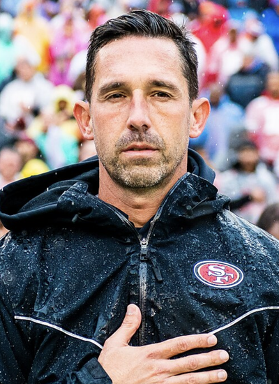 Close-up of 49ers head coach Kyle Shanahan wearing a rain-soaked black team jacket, hand over his heart, standing before a crowded stadium.