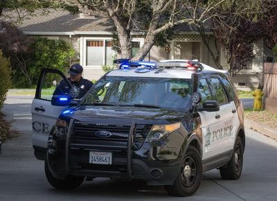 A Novato Police Department SUV with emergency lights activated, parked on a residential street as an officer stands by the open driver's door.