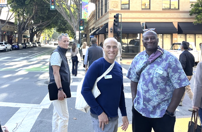 Three men wearing event name badges stand and smile on a sunny downtown street corner lined with shops, awnings, and pedestrians.