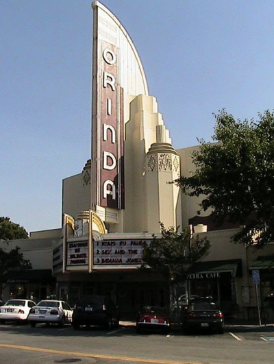 The art deco Orinda Theatre marquee towers over downtown Orinda's Theatre Square, with parked cars and Petra Café visible below.