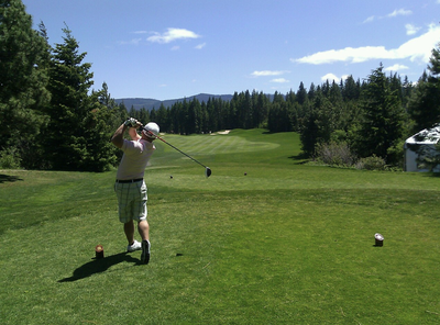 A golfer in a white cap and plaid shorts follows through on a tee shot, driver in hand, overlooking a tree-lined fairway with distant mountains.