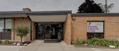 Entrance to a low brick YMCA building with a sign reading "Ensuring a Brighter Future," a covered walkway, and landscaping out front.