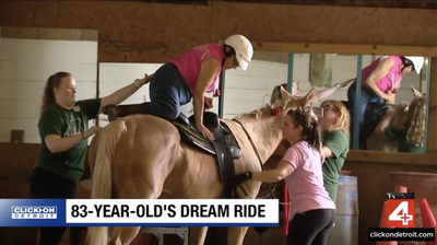 Karen Cooney, 83, wearing a pink shirt and helmet, is helped onto a horse by staff at Willowtree Stables, in a still from a WDIV Local 4 "ClickOnDetroit" news segment titled "83-Year-Old's Dream Ride."