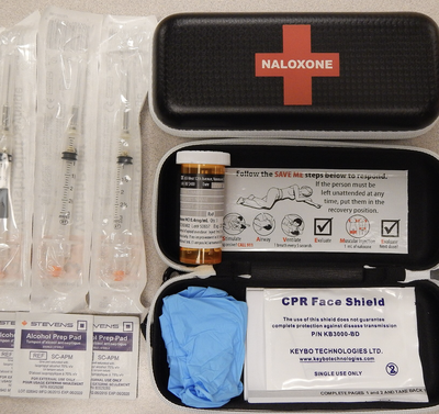A naloxone overdose-reversal kit containing syringes, a vial of naloxone, alcohol prep pads, gloves, and CPR instructions.