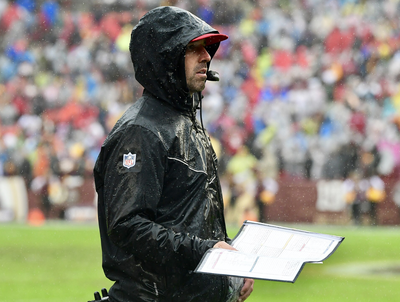 San Francisco 49ers head coach Kyle Shanahan stands on the sideline in the rain, wearing a hooded black rain jacket with an NFL patch, holding a play sheet.