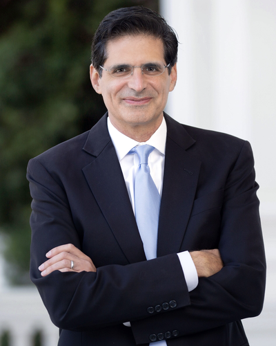 Portrait of State Sen. Josh Becker, a man with dark hair and glasses, wearing a dark suit and light blue tie, arms crossed, standing on a columned porch.