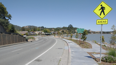 A curving road along a waterfront with a pedestrian-crossing warning sign in the foreground and hills across the water in the distance.