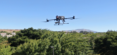 A San Rafael police drone hovers above treetops with hills and homes in the background.