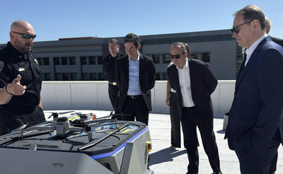 A bald police officer wearing sunglasses gestures toward a large drone unit on a rooftop while several men in suits look on during a daytime demonstration, with an office building in the background.