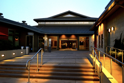 The Lafayette Library and Learning Center's entrance at dusk, with warm lighting illuminating the stone facade, steps, and railings, and the Bookmark Cafe visible to the left.