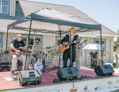 A band performs on an outdoor stage under a canopy tent at a past Tiburon Jewish Festival, with a guitarist in a black suit and cowboy hat singing at the mic.