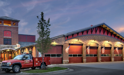 A Rochester Fire Department station at dusk, with a red fire truck parked out front and American flags mounted on the garage bay doors.