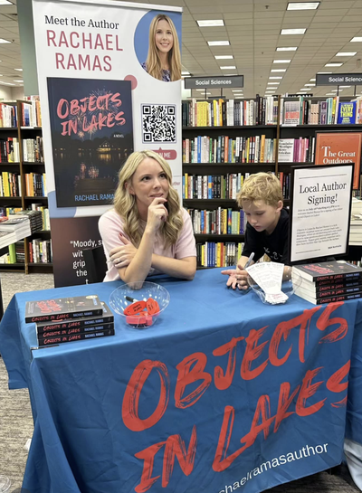 Author Rachael Ramas sits at a table signing a book for a boy, with a 'Meet the Author' poster and stacks of her novel 'Objects in Lakes' displayed at a Barnes & Noble store.