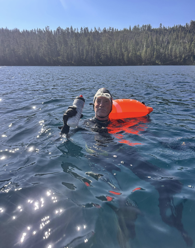 A swimmer wearing a wetsuit, cap, and goggles treads water in a lake, holding a water bottle, with an inflatable orange safety buoy attached to his waist and a pine forest shoreline in the background.