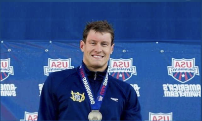 Peter Vanderkaay smiles on a podium wearing a USA Swimming team jacket and a silver medal, standing in front of a backdrop with repeated "USA Swimming" and "America's Swim Team" logos.