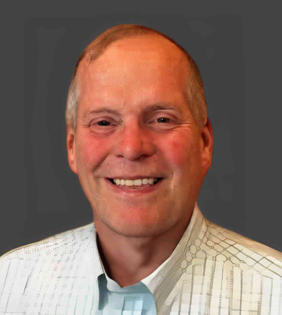 Headshot of Brent Johnston, a smiling man with short gray hair, wearing a light-colored patterned shirt against a dark gray background.