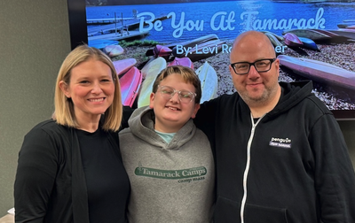 A woman, a young teenage boy wearing glasses and a Tamarack Camps sweatshirt, and a man pose together smiling in front of a screen displaying "Be You At Tamarack" with a lake and canoes pictured behind the text.