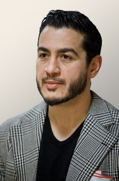 Abdul El-Sayed, director of Wayne County's Department of Health, Human, and Veterans Services, in a black-and-white houndstooth blazer and black v-neck shirt, looking off to the side against a plain background.