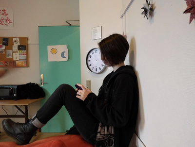 A teenager sits on the floor against a wall, looking down at a phone.