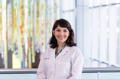 Dr. Isabella Comsa, DO, wearing a white coat, smiling in a hospital lobby.